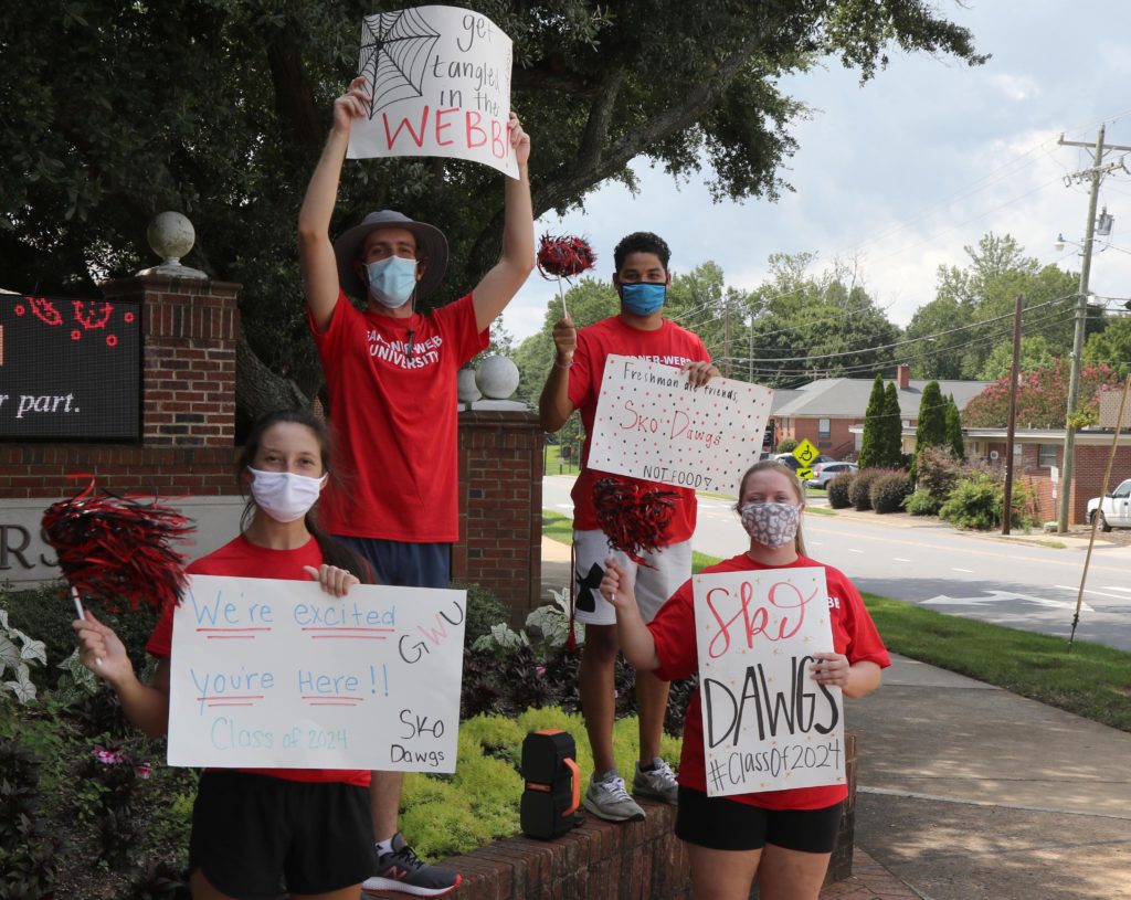 Four students hold up signs as they stand on the street corner welcoming new students to Gardner-Webb
