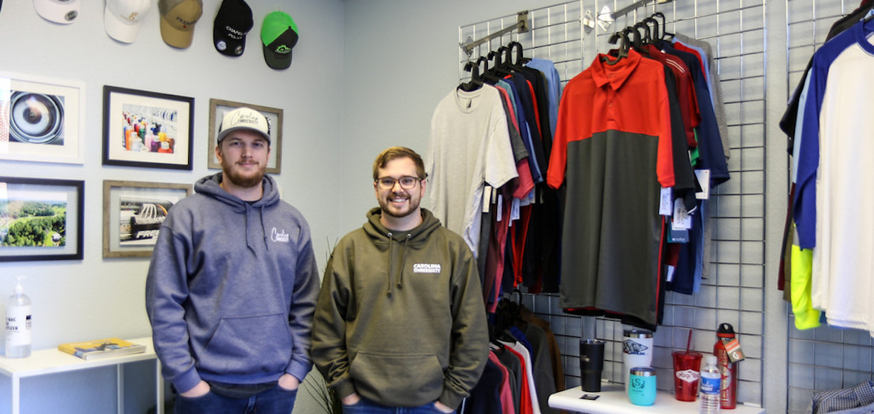 Cam, left, and Bryton Mitchell are brothers and GWU alumni who recently started their own business. They are posing in their shop, with shirts and hats and other items that people can have personalized.