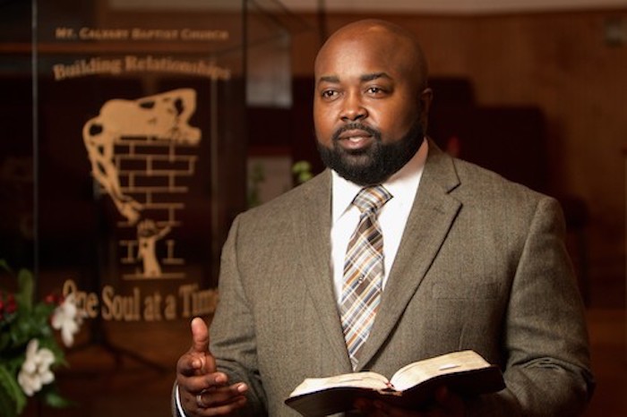 A photo of Rev. Dr. M. Lamont Littlejohn holding a Bible and standing in his church sanctuary.