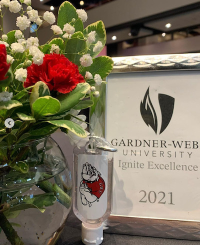 The table display at the Ignite Excellence Scholarship Interviews, featuring a floral arrangement with red flowers, a GWU hand sanitizer with the bulldog logo and a sign that reads Gardner-Webb University Ignite Excellence 2021