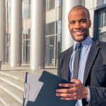 Handsome young Afro American businessman in classic suit holding a folder, looking at camera and smiling, standing outside the office building