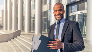 Handsome young Afro American businessman in classic suit holding a folder, looking at camera and smiling, standing outside the office building