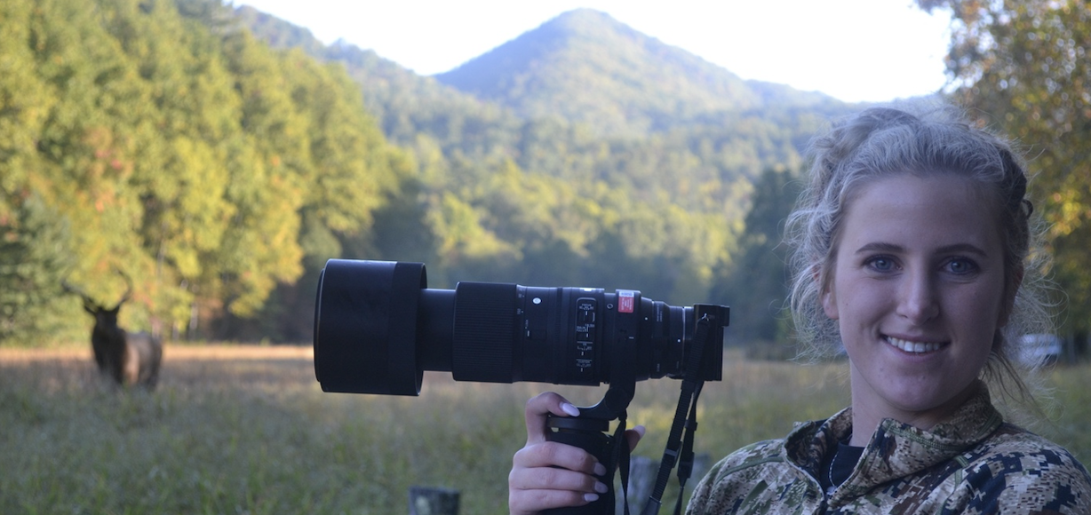 A student poses in the Cataloochee Valley with a camera and an elk in the background